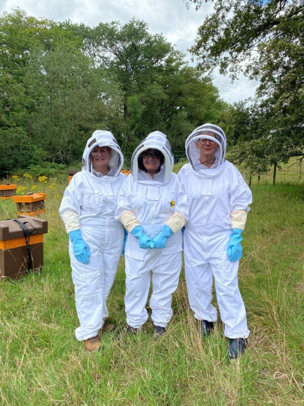 Three ladies in bee suits smiling in front of bee hives